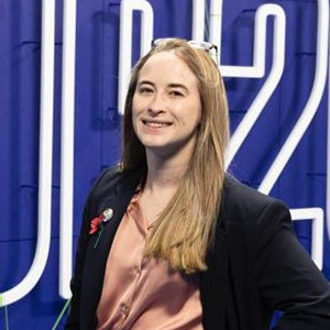 Isabel Webb wearing a dark blazer and pink shirt standing in front of a blue illuminated display with large lettering.