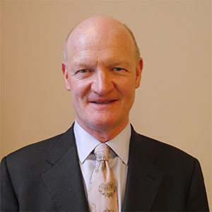 Professional headshot of a person wearing a dark business suit, light shirt and patterned tie against a plain beige background.